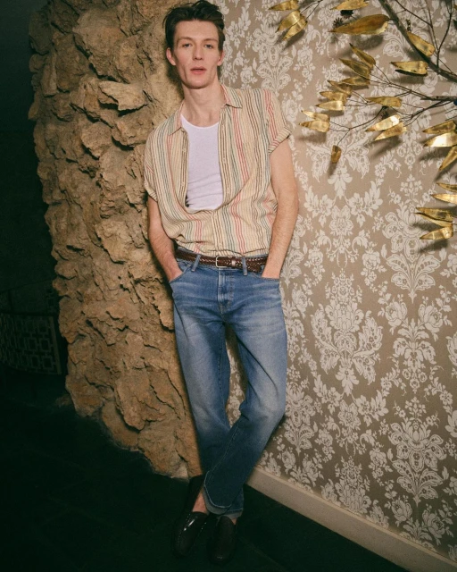 Man in jeans and striped shirt leaning against patterned wall and rock texture, casual style, indoor setting, medium shot.