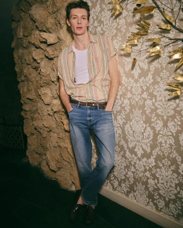 Man in jeans and striped shirt leaning against patterned wall and rock texture, casual style, indoor setting, medium shot.