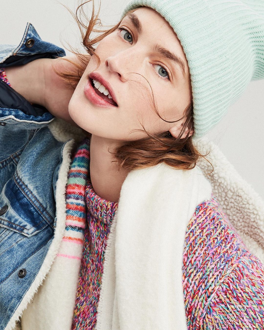 Close-up of a young adult woman in a knit hat, colorfully speckled sweater, and denim jacket, looking at the camera with a slight smile.