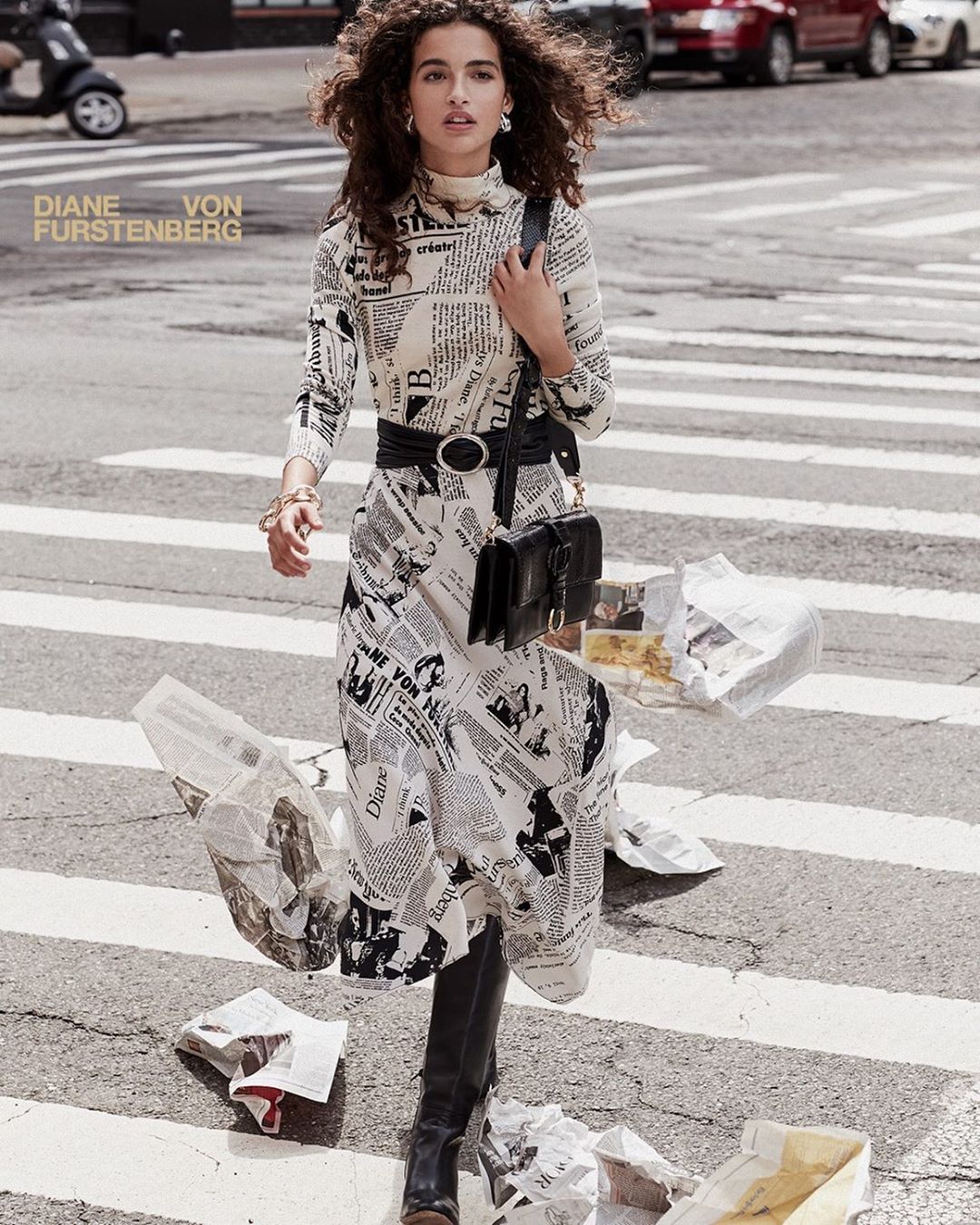 A woman walks on a crosswalk wearing a white newspaper-print dress, black boots and purse, with cars and a scooter in the background.