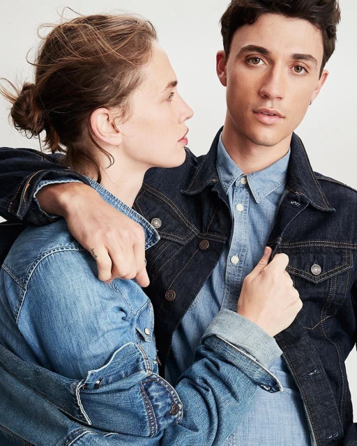 A fashionable couple in denim jackets poses indoors against a white background, showcasing stylish casual wear in a studio-like setting.