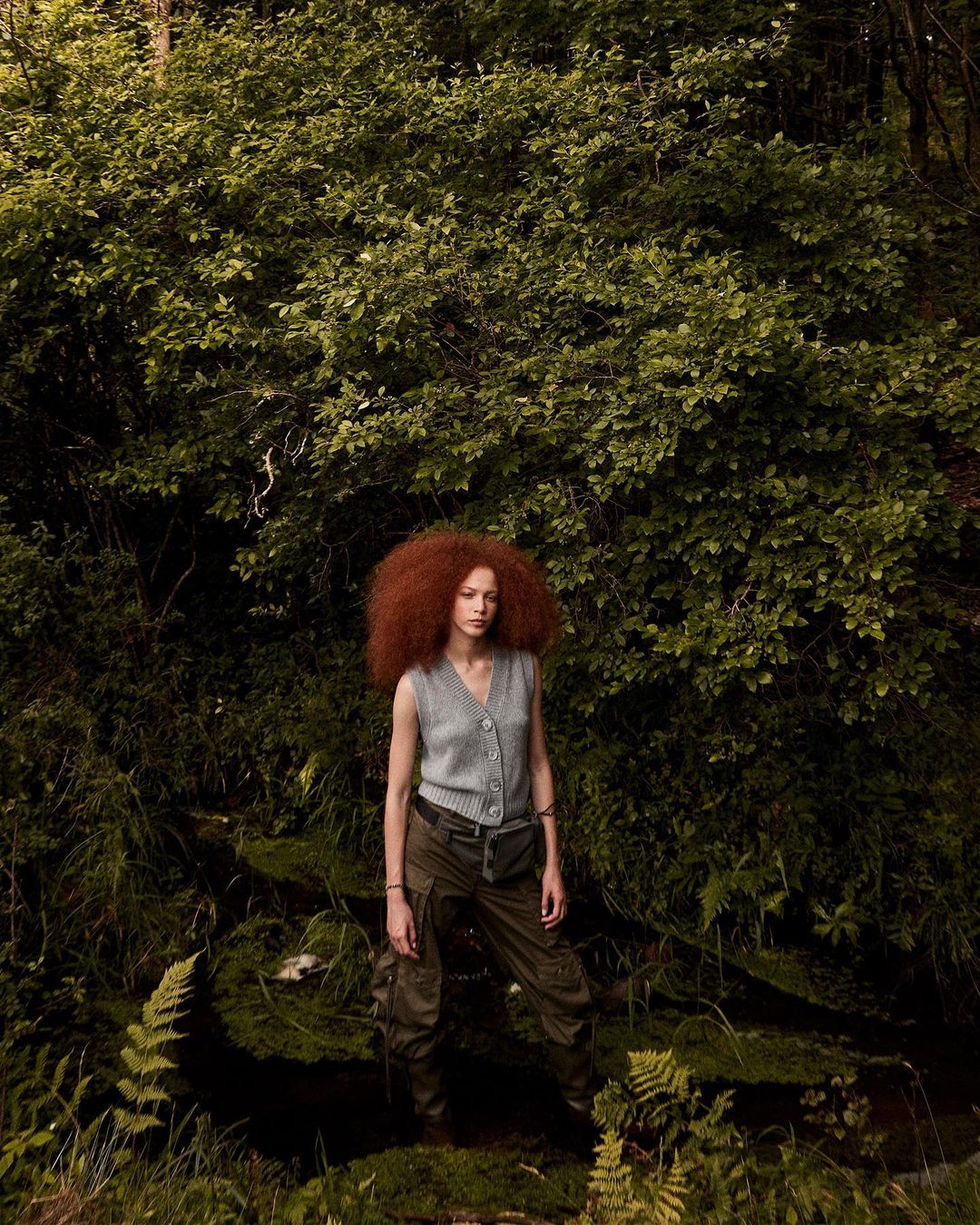 Adult woman with red hair poses in a sleeveless top and dark pants in a forest setting with green foliage and dappled light.