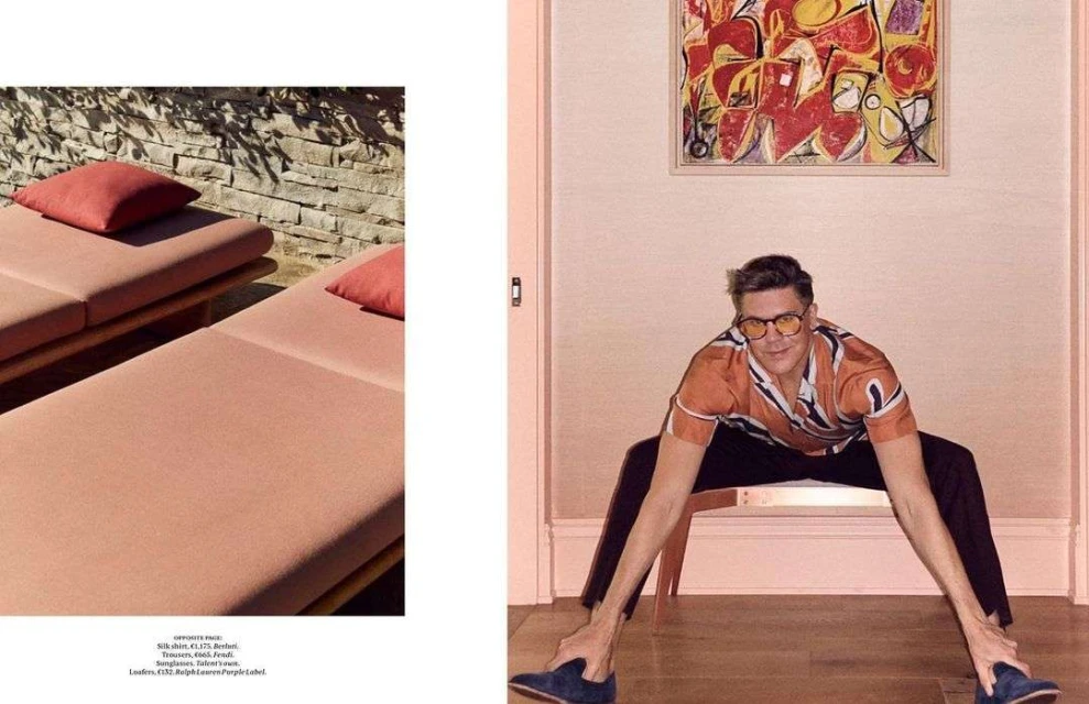Indoor fashion shot of a man in an orange pattern shirt and black pants sitting low with stretched legs and feet touching the floor.