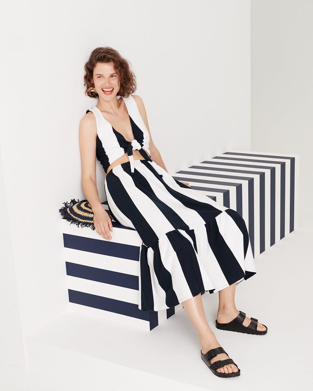 A woman in a striped two-piece set poses sitting on a striped cube in a light studio with a straw hat next to her and black sandals.