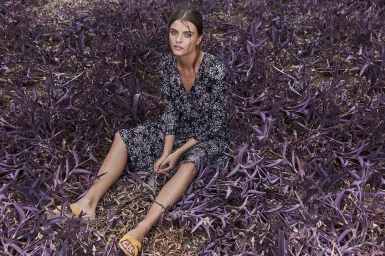 A woman seated amidst a field of vibrant purple foliage wearing a patterned dark dress and yellow sandals.