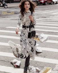 Model in newspaper print dress and black boots walking across a city crosswalk with scattered papers.