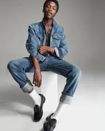 Young Black man in denim jacket and jeans seated on a white block against a grey studio backdrop