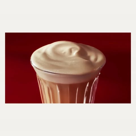 Frothy beverage in a clear glass with a red background. The drink is topped with a thick layer of creamy foam.