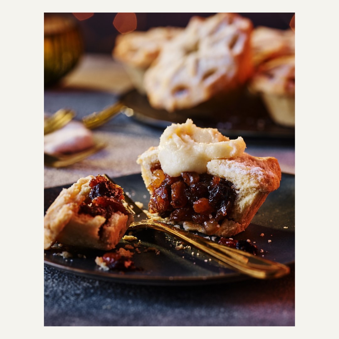 Close-up of a cut-open mince pie with cream on a plate, showcasing its rich fruit filling, powdered sugar dusting, and golden fork.