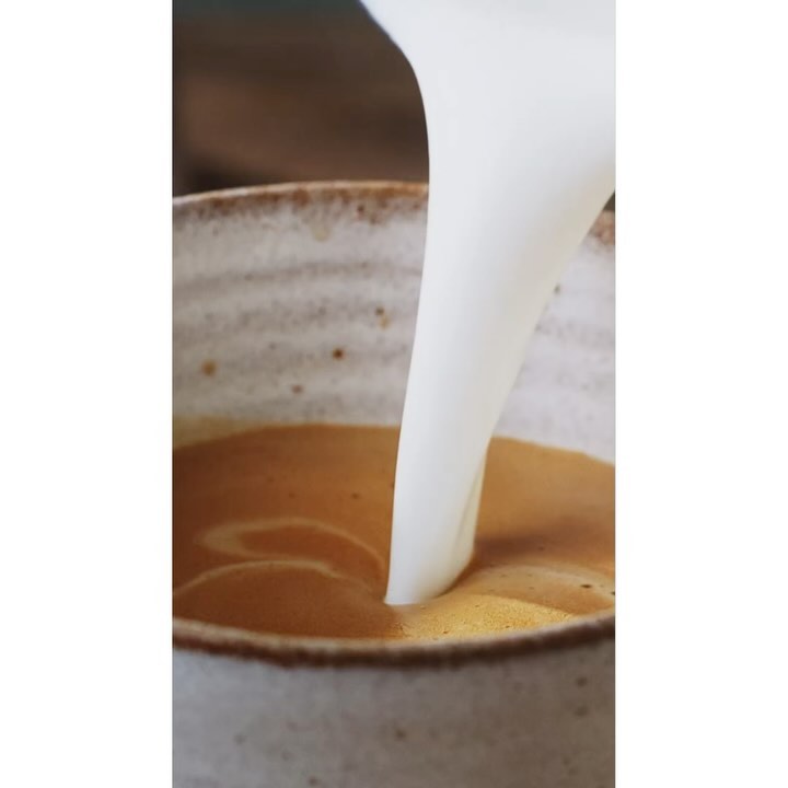 Close-up of milk being poured into a cup of coffee, creating a creamy swirl in a speckled ceramic mug. Warm, inviting beverage.