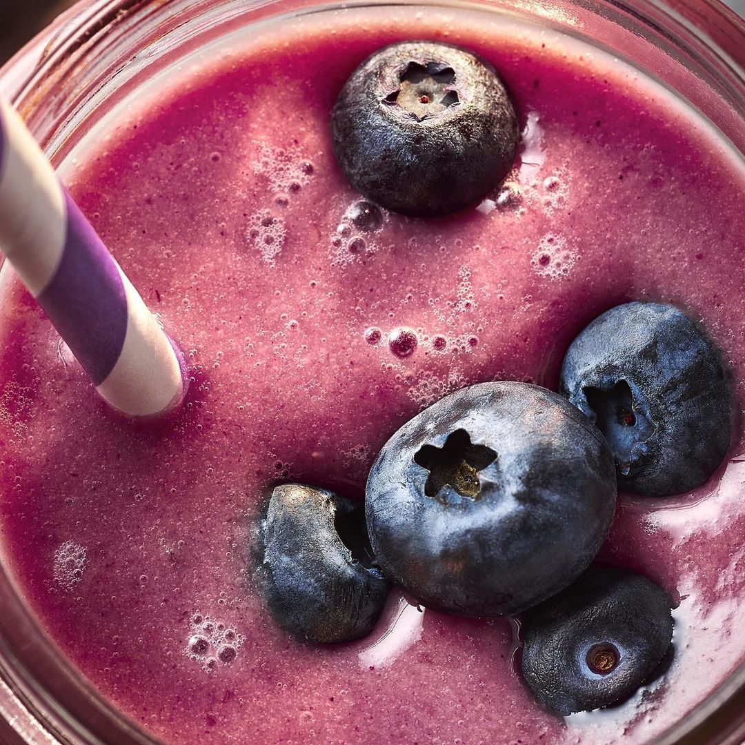 Close-up of a blueberry smoothie in a glass with a straw. The smoothie has a purple hue and is topped with several fresh blueberries.