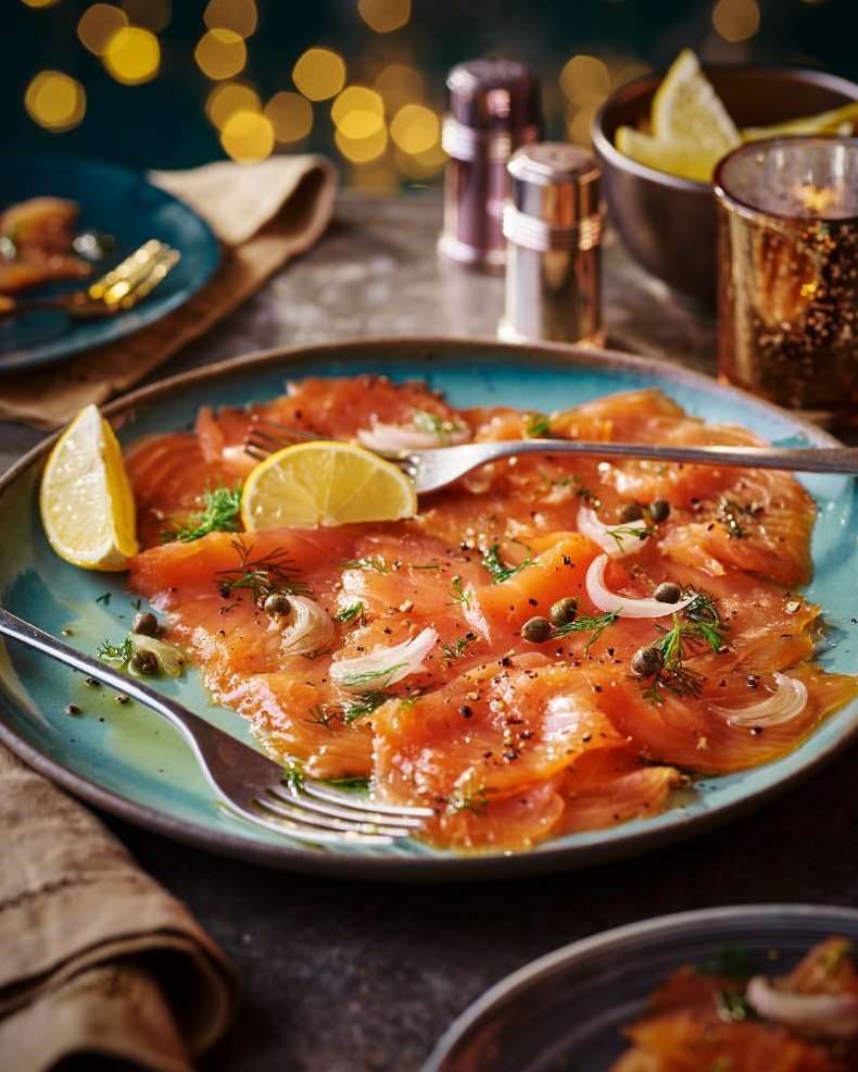 Overhead shot of smoked salmon on a blue plate, garnished with lemon, dill, capers, and sliced onion; flatware on the side, moody lighting.