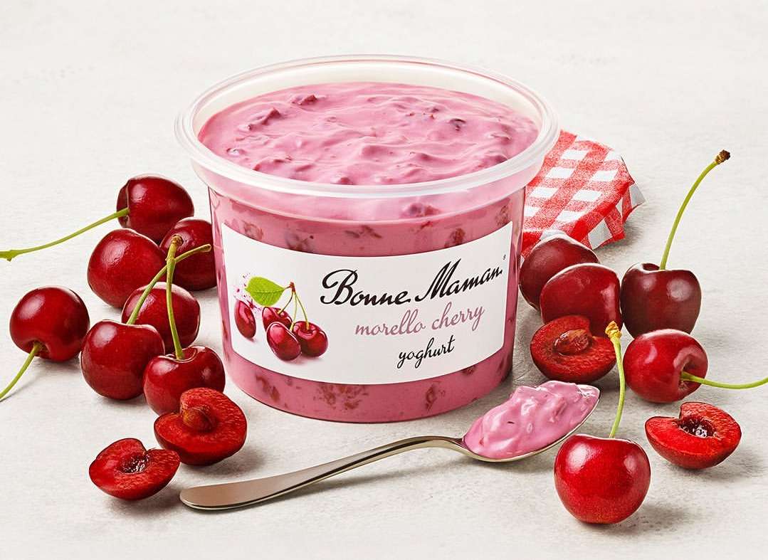 Cherry yogurt in a clear container, surrounded by fresh cherries and a silver spoon with yogurt, against a white background.