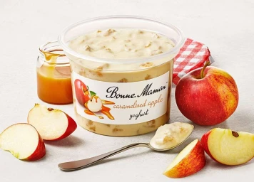 Plastic tub of caramelised apple yoghurt surrounded by fresh red apples, apple slices, a jar of caramel, and a spoon.