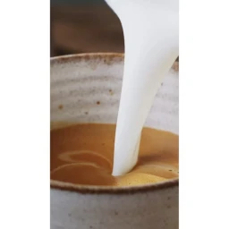 White milk pouring into a ceramic mug filled with brown coffee, captured in a close-up shot by Colin Campbell.