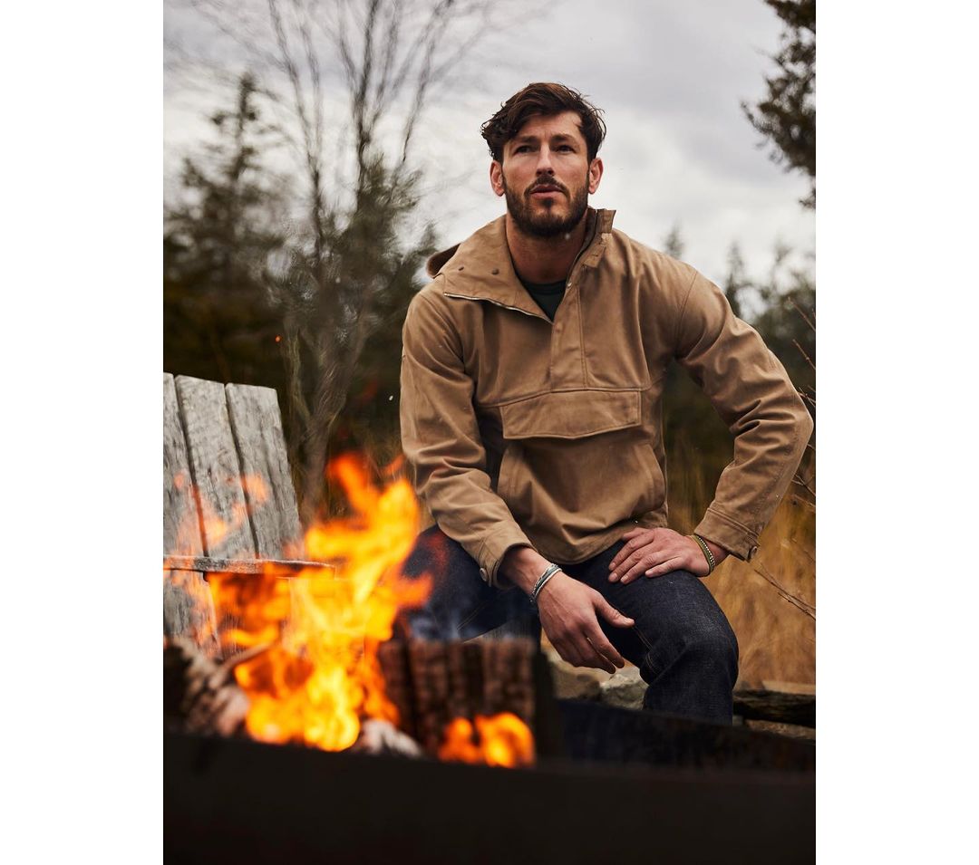 Adult male wearing tan jacket sits outdoors by campfire in rural setting. Outdoor fashion shot featuring fire, wood, and neutral tones.