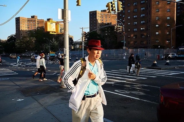 Man in a hat crosses a street in an urban setting with apartment buildings, traffic signals, and other people walking nearby on a sunny day.