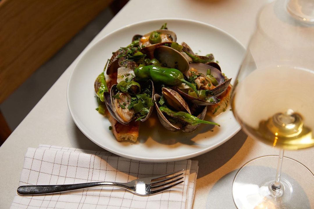 Overhead shot of a plate of clams and green peppers with a fork and wine glass on a white table in a restaurant setting.