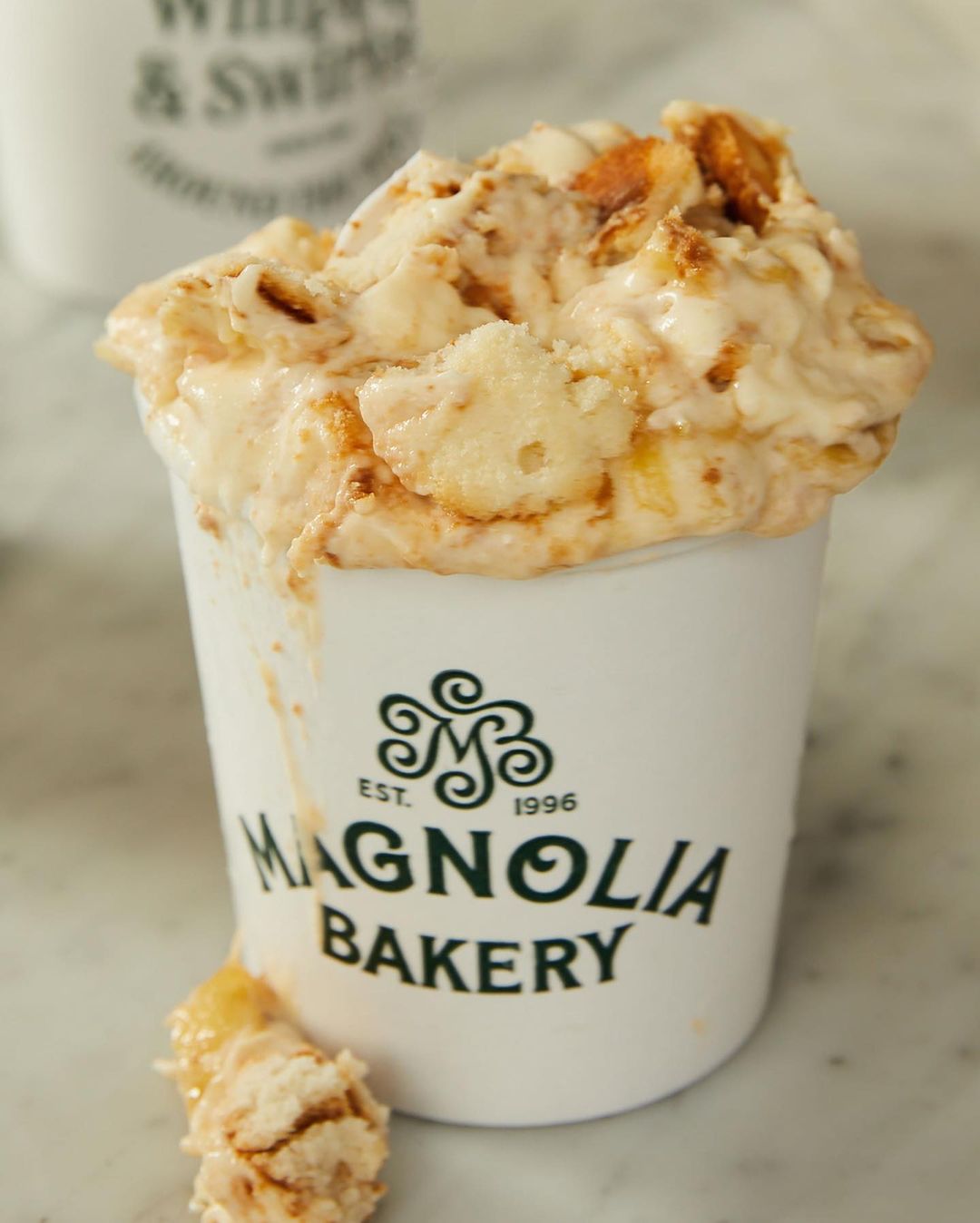 Magnolia Bakery banana pudding in a cup with logo on marble surface, rich in creamy texture and golden hues, food photography.