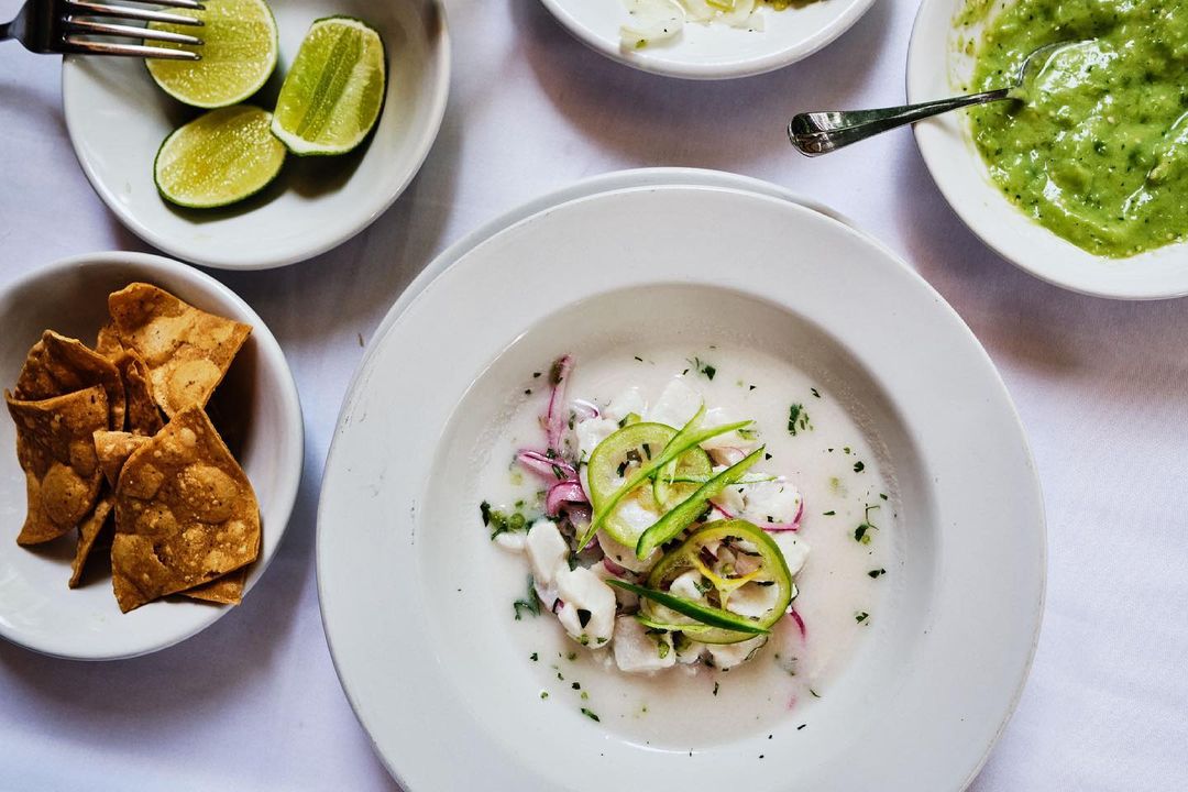 Overhead shot of a plate of ceviche with lime wedges, tortilla chips, and salsa verde on a white tablecloth, highlighting food photography.