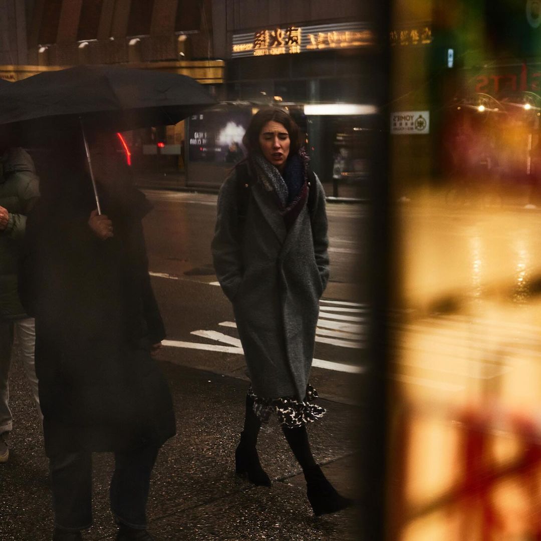 Woman in a grey coat walks on a city street at night. She is accompanied by a person with an umbrella in rainy weather.