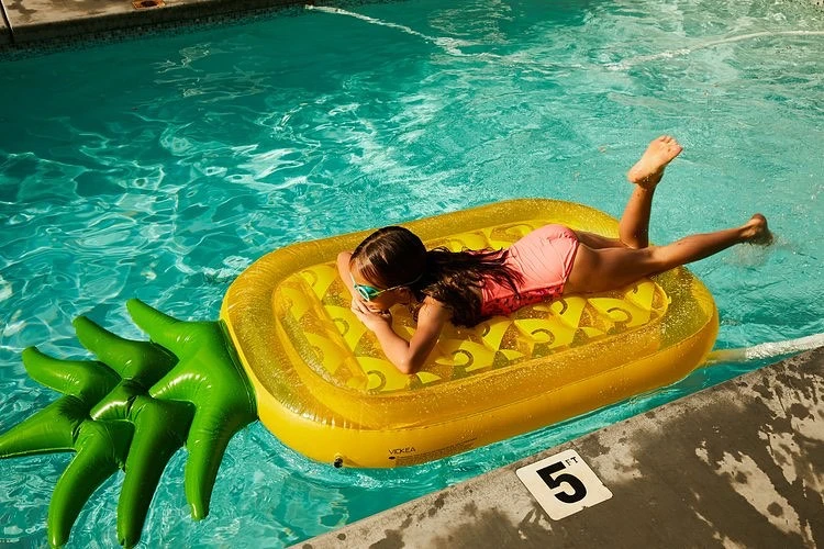 A young girl in a pink bathing suit lies on a yellow pineapple float in a swimming pool on a sunny day enjoying a summer vacation.