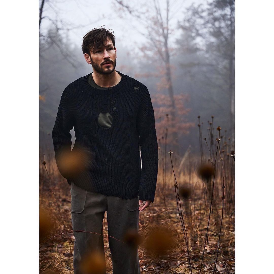 Adult man in a black sweater and khaki pants stands in a misty, wooded landscape in a medium shot with a shallow depth of field.