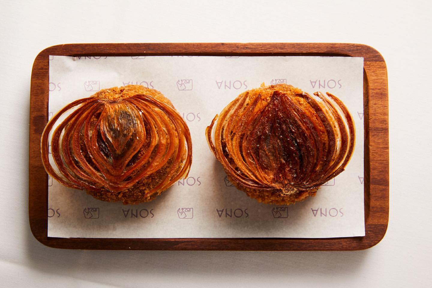 Two blooming onion appetizers on a wooden serving board, displayed on white paper and a light background in a top-down product shot.