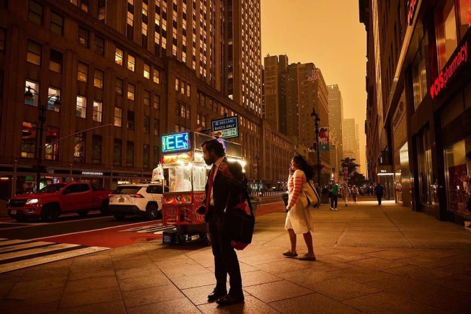 Urban street scene with pedestrians walking past food cart, cars, and tall buildings under a warm, orange-toned sky in New York City.