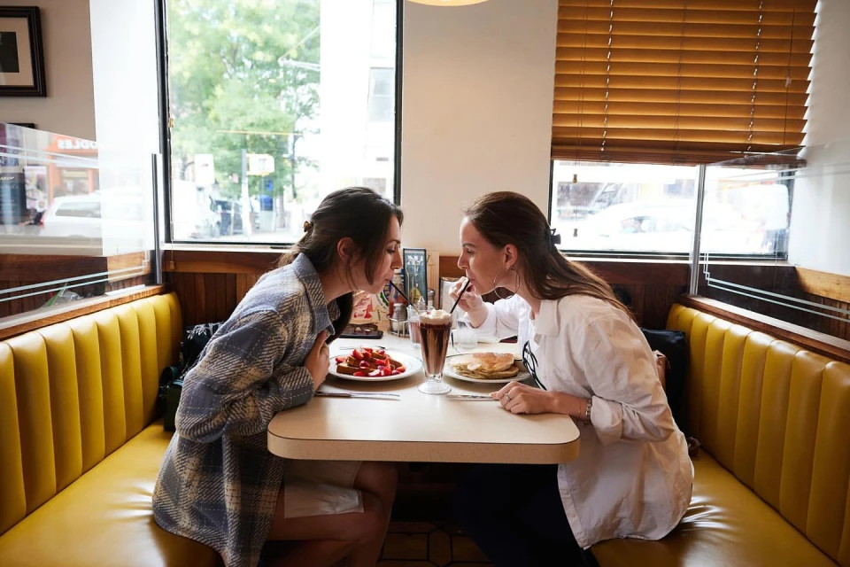 Two adult women sit in a diner booth, sharing a drink with two straws. Plates of food sit on the table in front of them.