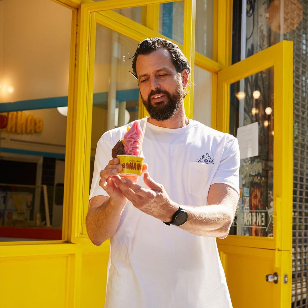 Man holding a yellow cup of pink and brown frozen dessert outdoors in front of a yellow building with glass windows and white trim.