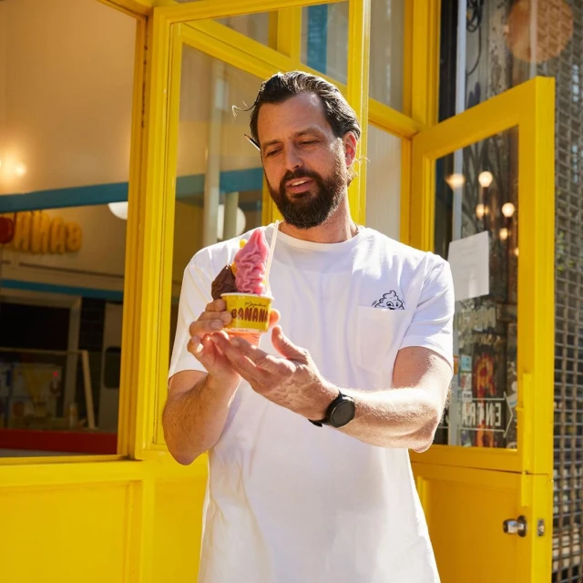 Man holding a yellow cup of pink and brown frozen dessert outdoors in front of a yellow building with glass windows and white trim.