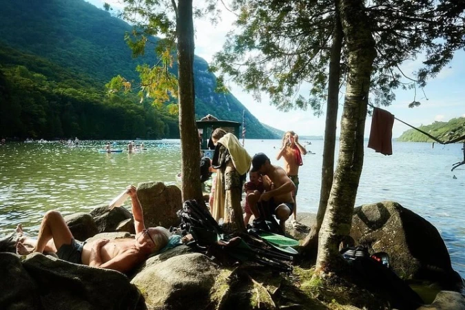 Adults and children relax by a lake on a sunny day with forested mountains in the distance. Some people are swimming in the water.