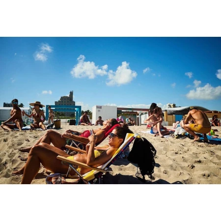 Several people relaxing on a sandy beach with lounge chairs, and some standing or sitting, under a clear, bright sky on a sunny day.