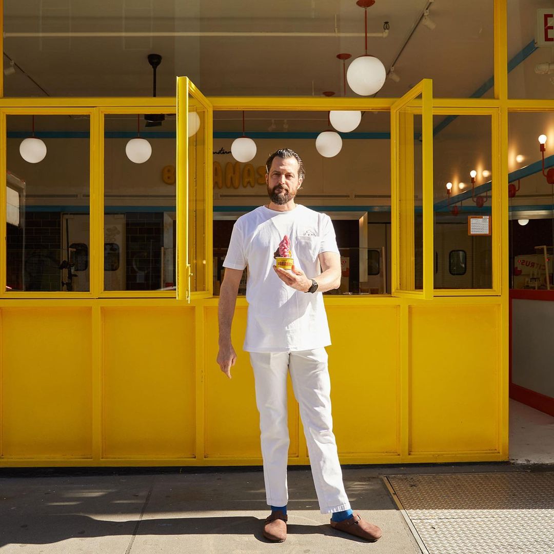 Adult man holding a pink-colored ice cream cone in front of a vibrant yellow building with hanging spherical lights.