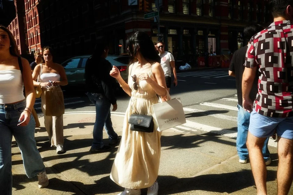 A woman in a dress walks along a sunny city street carrying a shopping bag, accompanied by other pedestrians on a bright day.
