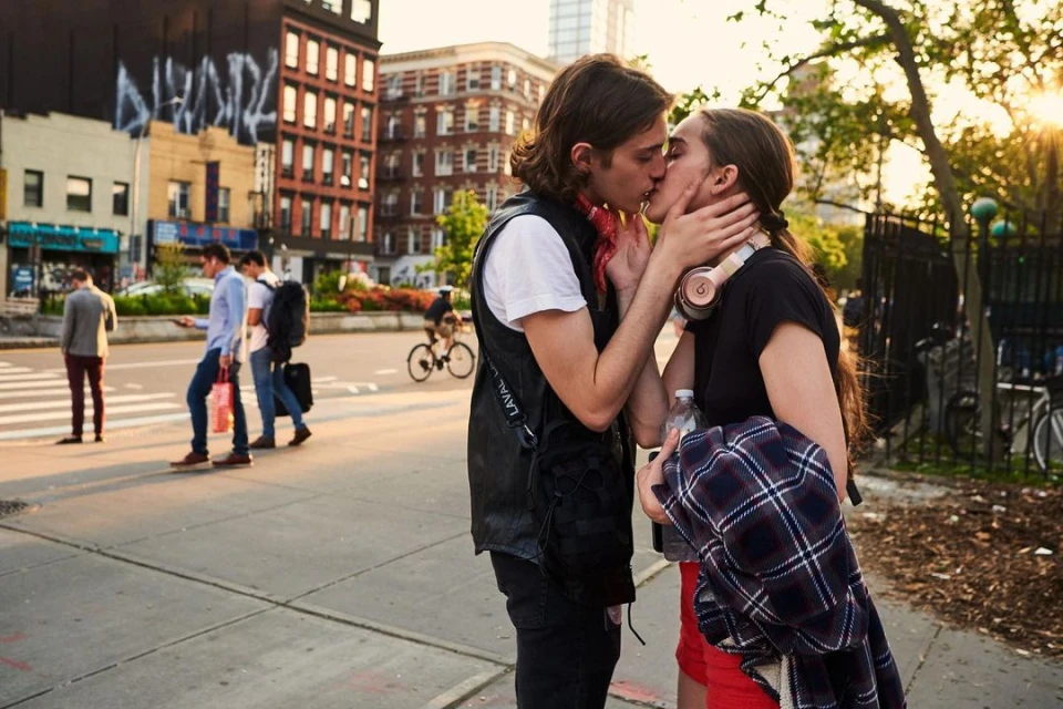 Outdoor shot of a kissing couple in an urban park, with pedestrians and buildings in the background during golden hour.