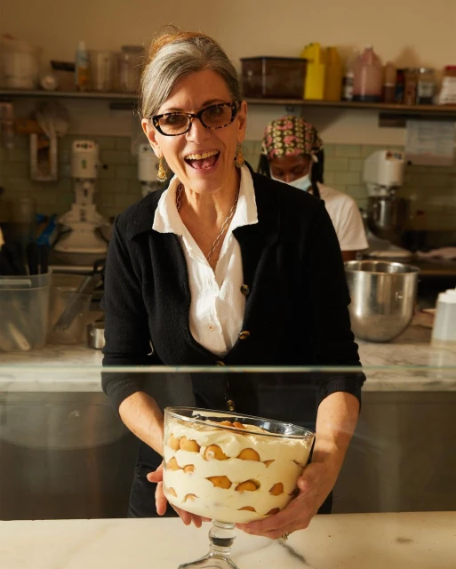 Smiling woman in kitchen holding banana pudding dessert in a clear trifle bowl. Commercial kitchen setting with another person visible.