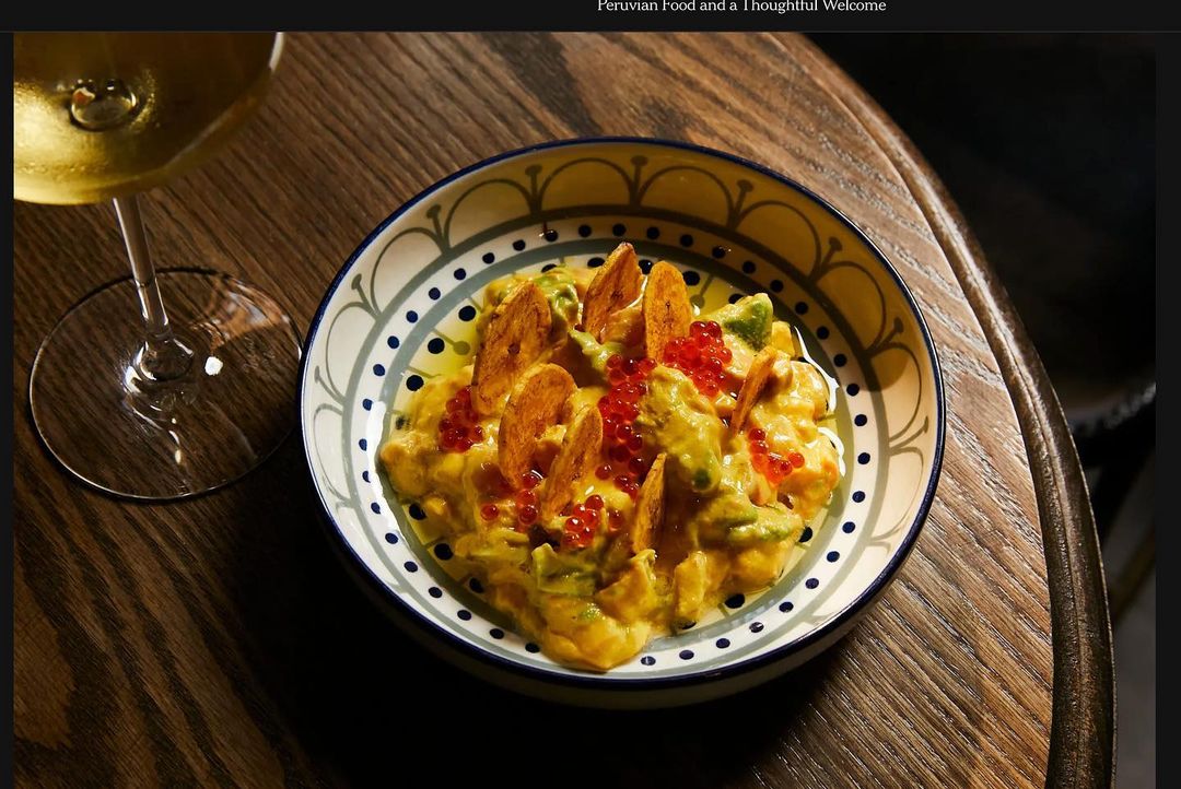 Overhead shot of Peruvian ceviche with fish, avocado, chili peppers, and fried plantains in a colorful bowl on a wooden table next to wine.
