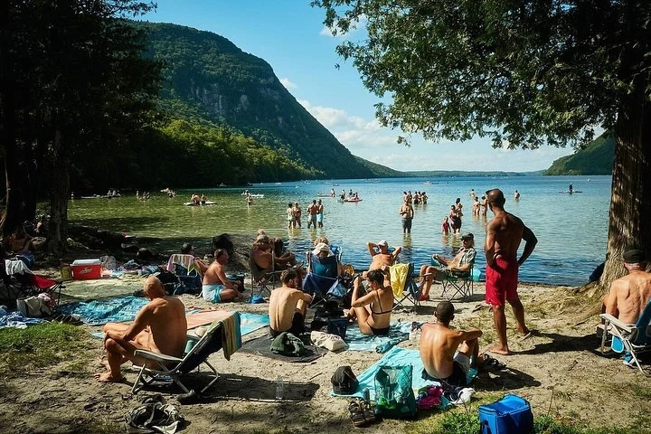 Daytime view of a sandy lakeside beach with numerous adults wading in the water and relaxing on the shore in folding chairs.