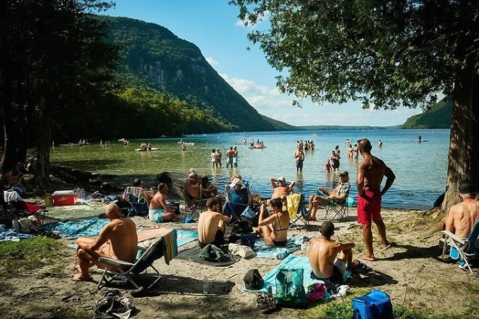 Daytime view of a sandy lakeside beach with numerous adults wading in the water and relaxing on the shore in folding chairs.