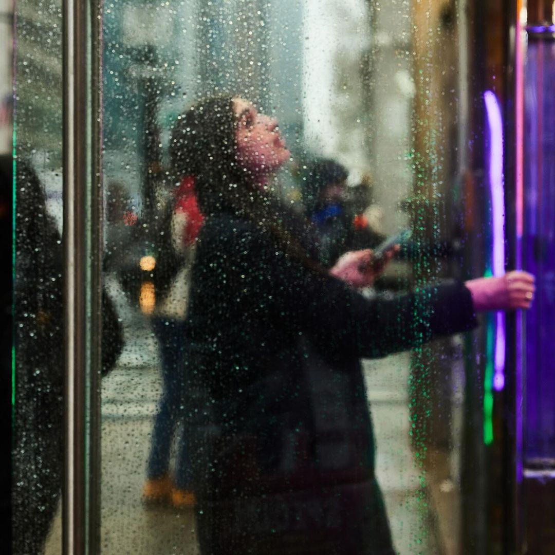 A woman with dark hair looks upward while standing behind a rain-covered glass door in an urban setting at night.