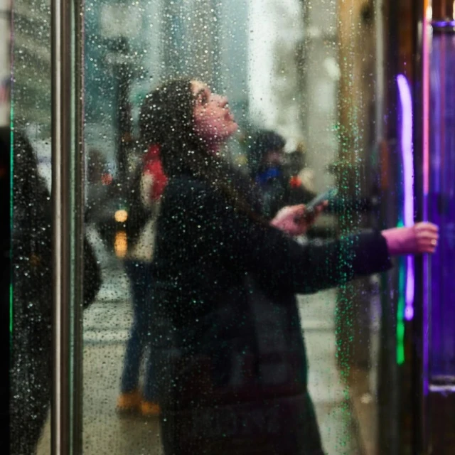 A woman with dark hair looks upward while standing behind a rain-covered glass door in an urban setting at night.