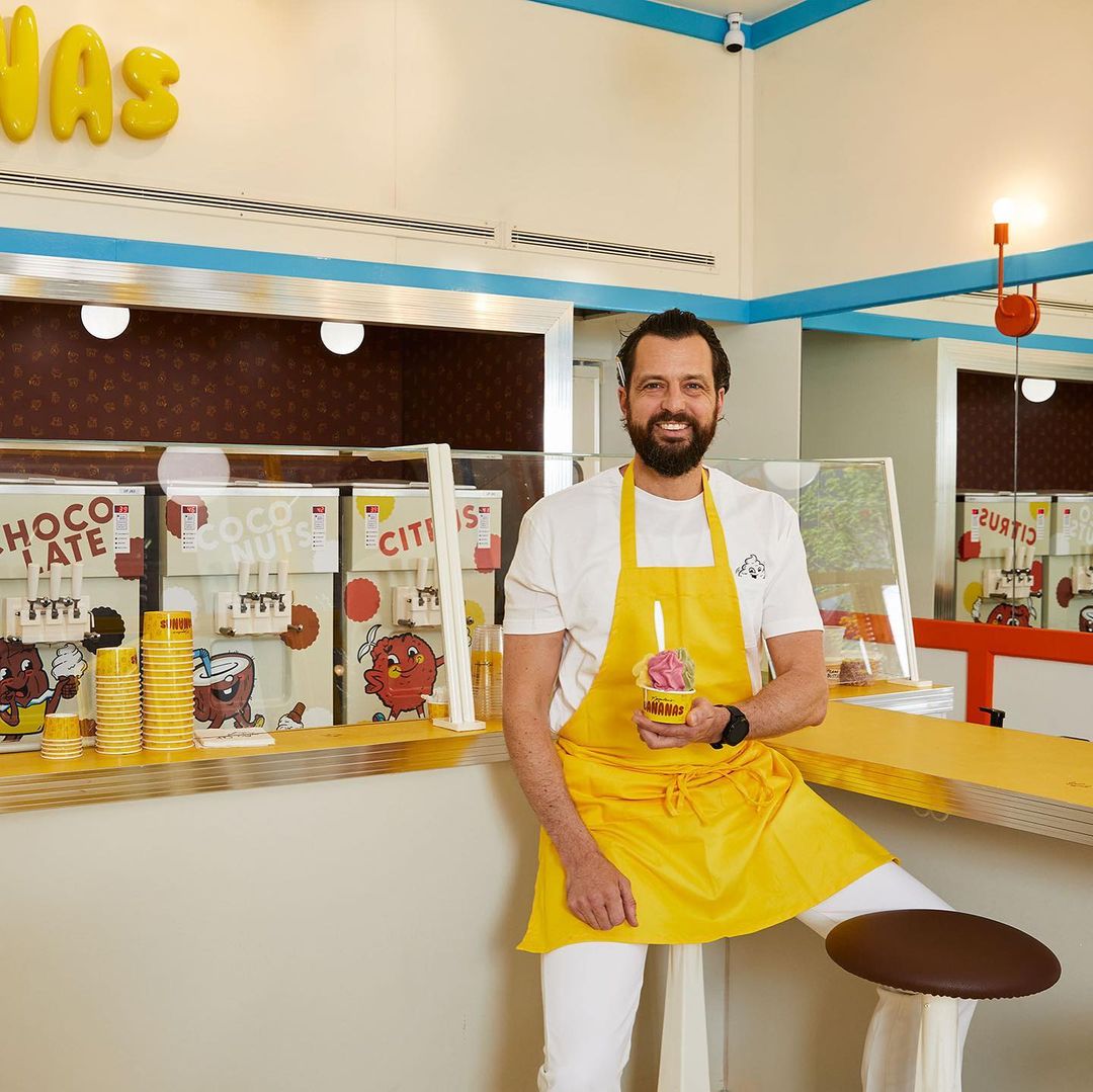 Bearded man wearing a yellow apron holds a cup of colorful ice cream in a bright, retro-styled dessert shop with vintage decor.