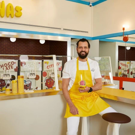 Bearded man wearing a yellow apron holds a cup of colorful ice cream in a bright, retro-styled dessert shop with vintage decor.