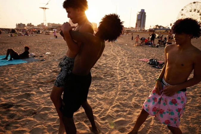 Three shirtless young men on a sandy beach during golden hour, with a crowd, buildings and a tall ride in the background.