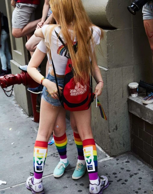 Two people stand wearing rainbow and LOVE-themed socks with a red backpack in an outdoor urban setting.