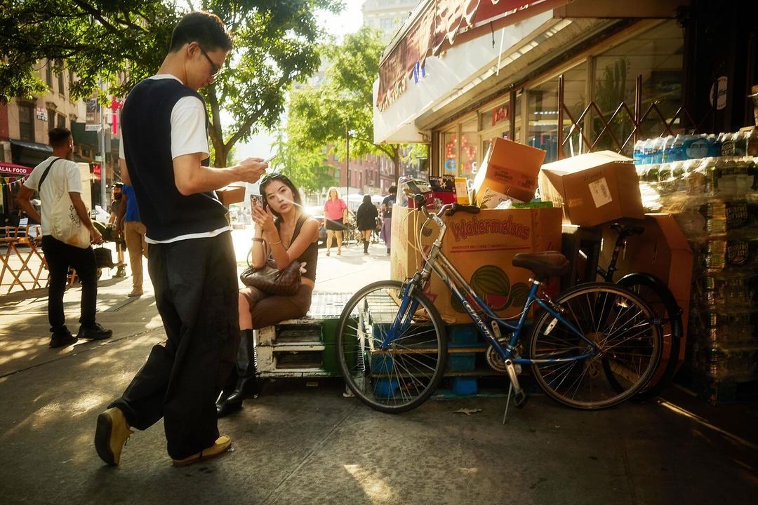 Man and woman standing on a New York sidewalk near stacked cardboard boxes and a blue bicycle, photographed by Colin Clark.