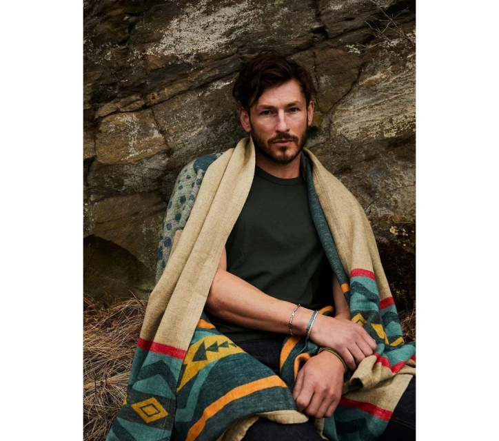 Man wrapped in blanket against rock wall. Male model poses in dark green shirt, colorful blanket, rocky outdoor setting.