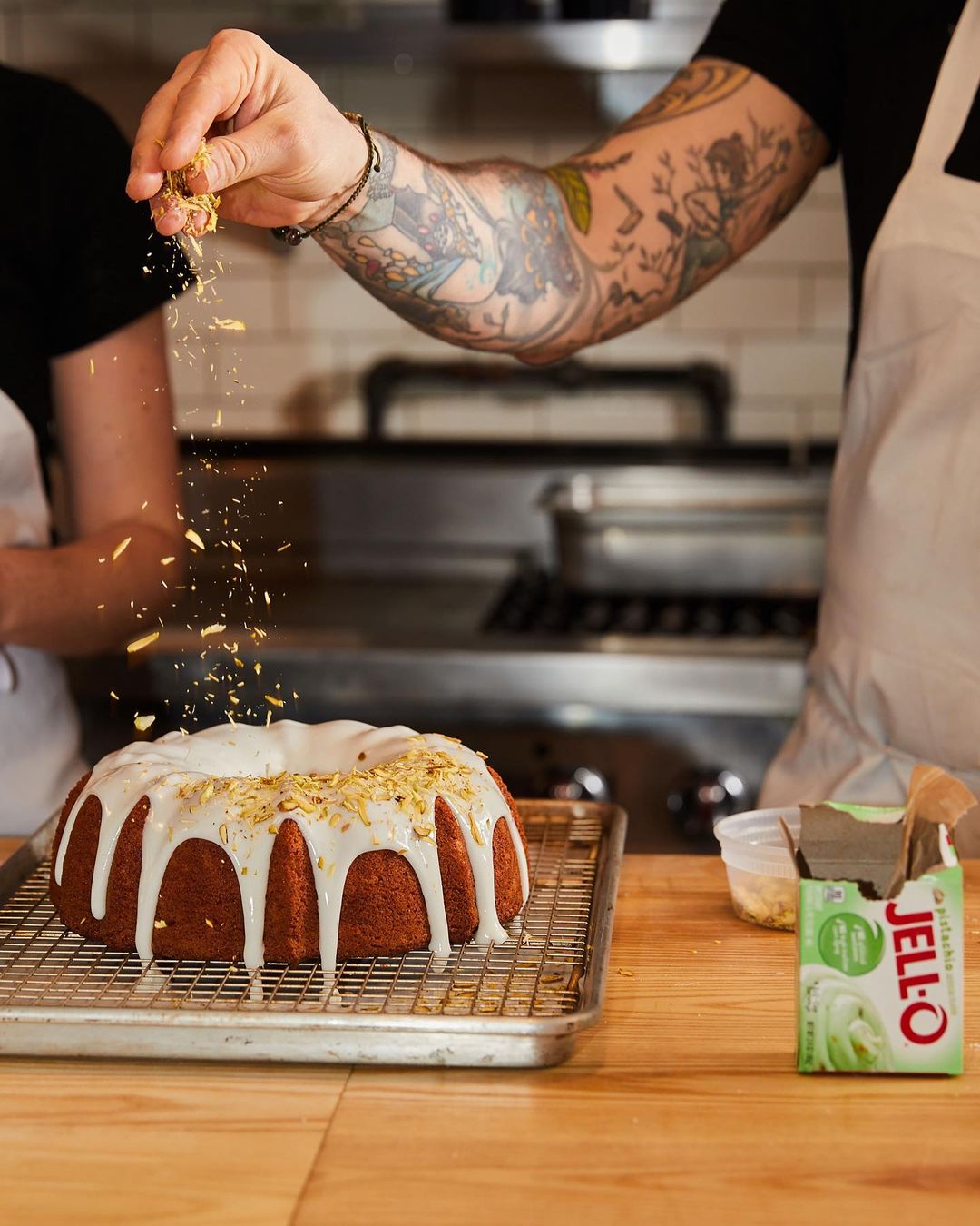 Ring cake on a metal cooling rack being decorated by a tattooed arm with sprinkles with Jell-O pudding mix on a wood table.